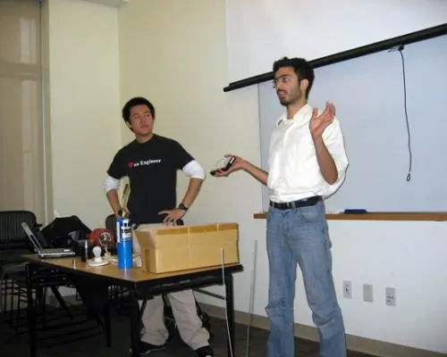 Two people stand behind a table at the front of a classroom while one of them addresses the audience.